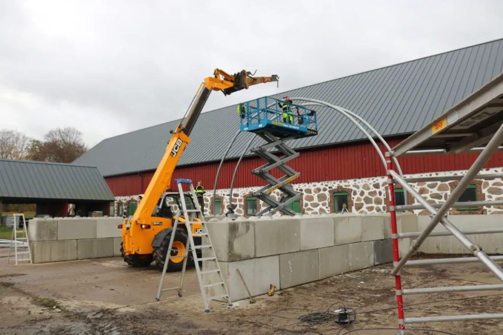 Montering av tälthall på betongblock vid en ladugård, med skylift och teleskoplastare